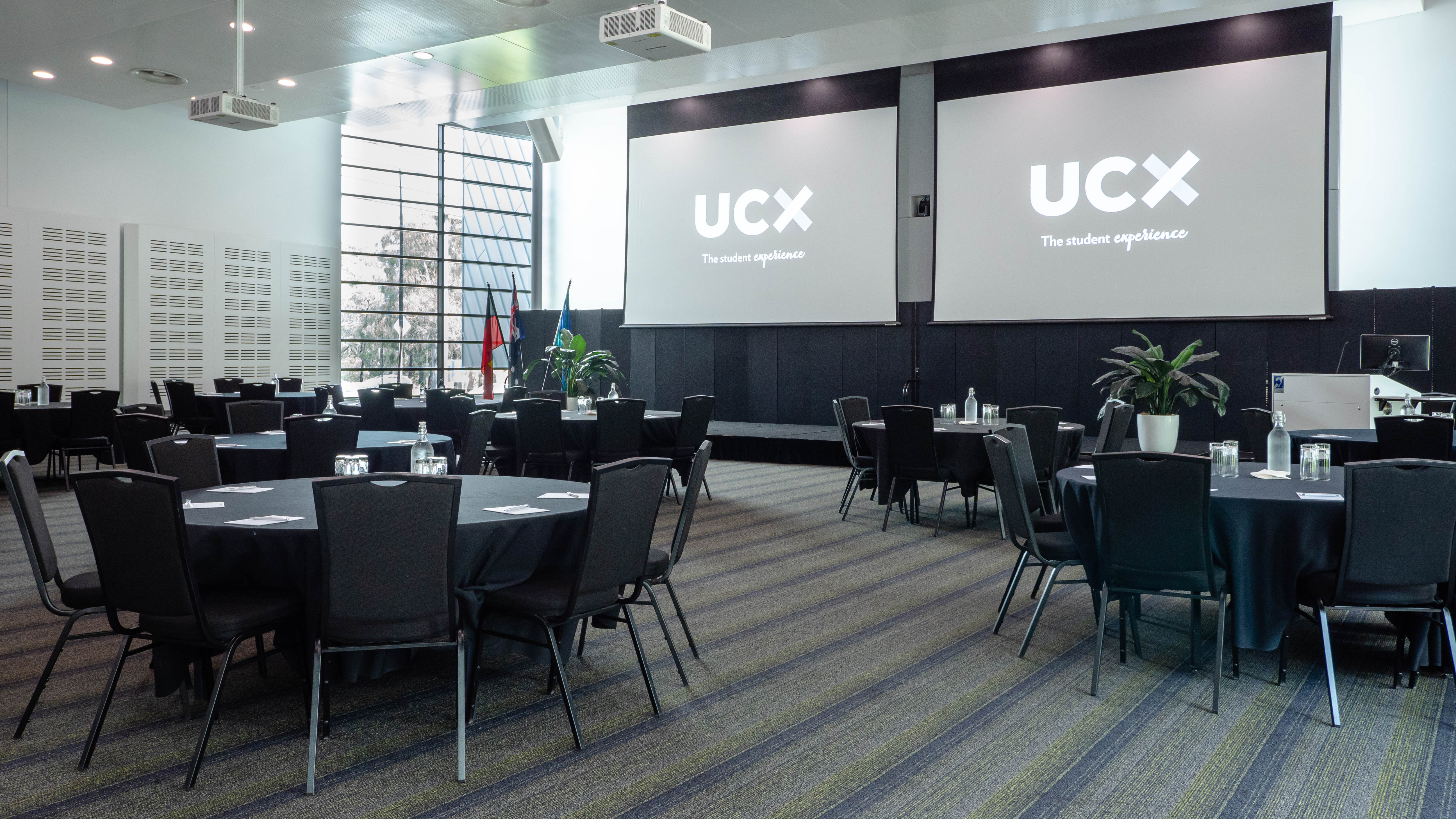 Multiple round tables with chairs around them in a large room. Each chair has a notepad and glass in front of it. The UCX logo is displayed on projector screens at the front of the room