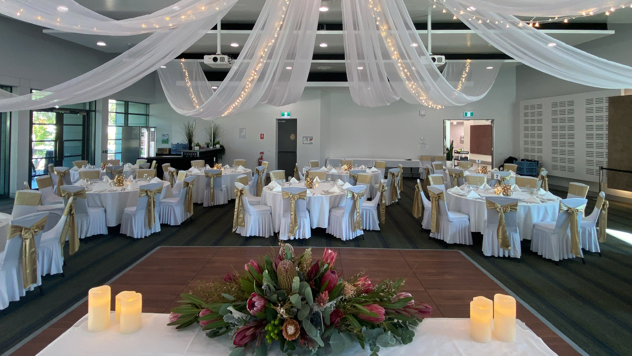 The Ann Harding Conference Centre set up for a wedding. There are multiple round tables with floral centrepieces and candles. Lighting and tulle is draped from the ceiling. 