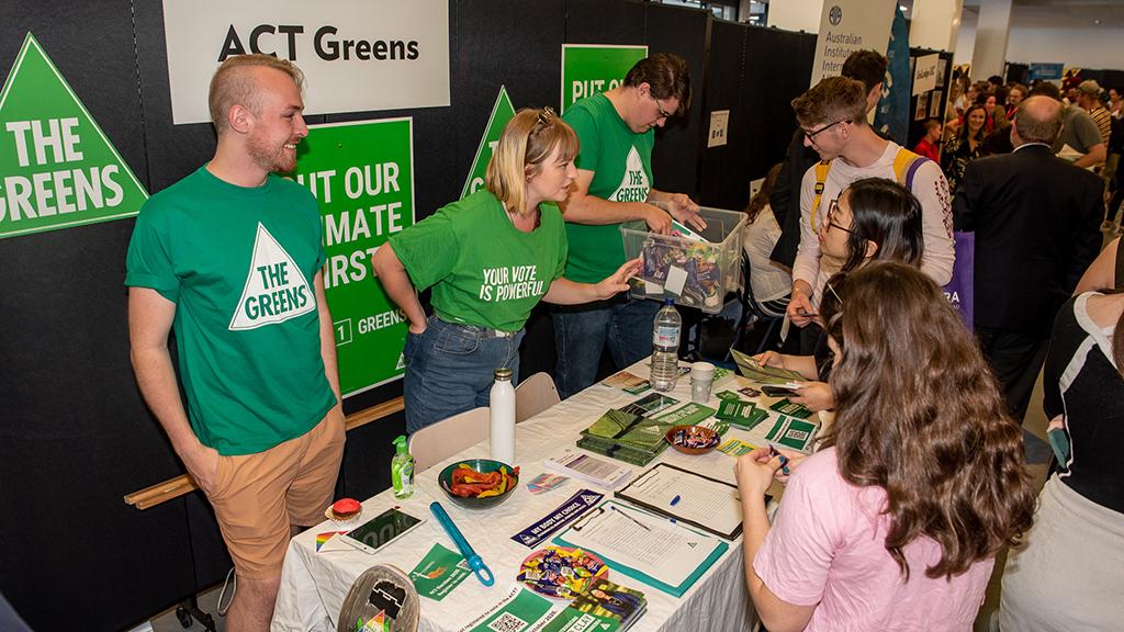 Three members of the UC Greens social club standing behind a table talking to a group of interested students.