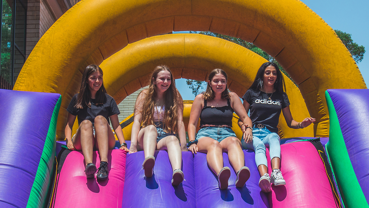 Four female students with apprehensive expressions at the top a giant inflatable slide about to slide down.