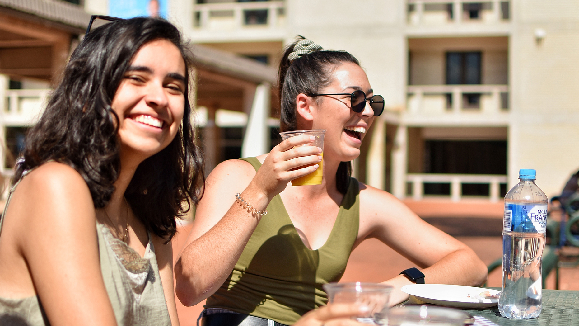 Two female students smiling and laughing while sitting at an outdoor table on the UC Concourse. The student on the right is holding a cup of juice after a free breakfast event in O-Week.