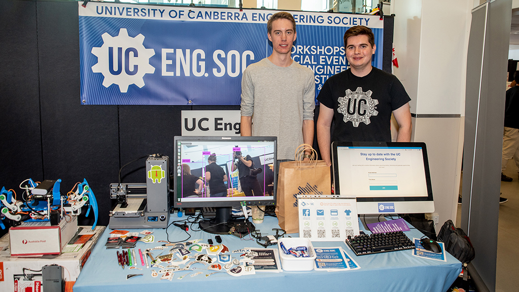 Two male students from the UC Engineering Society behind a table at market day. On the table are robots, information and videos playing on a monitor.