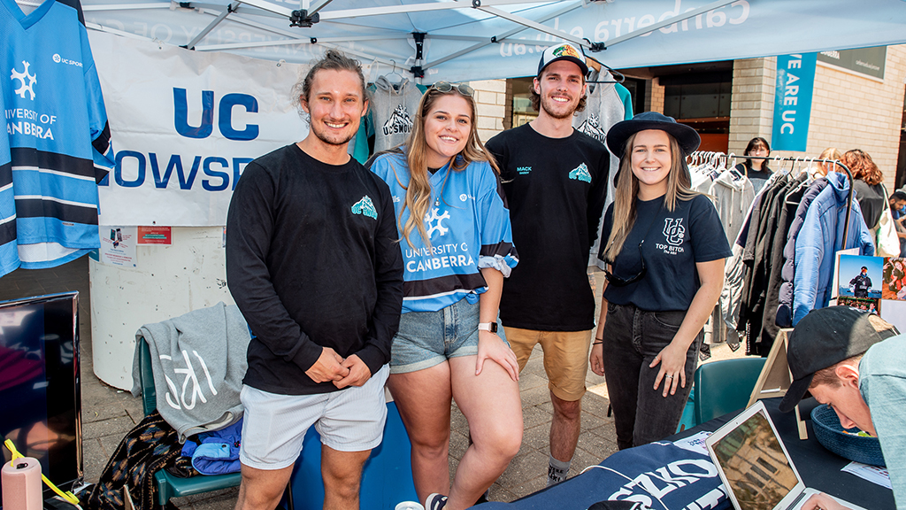 Four people standing under a marquee showcasing information and merchandise for the UC Snowsports Club outside.
