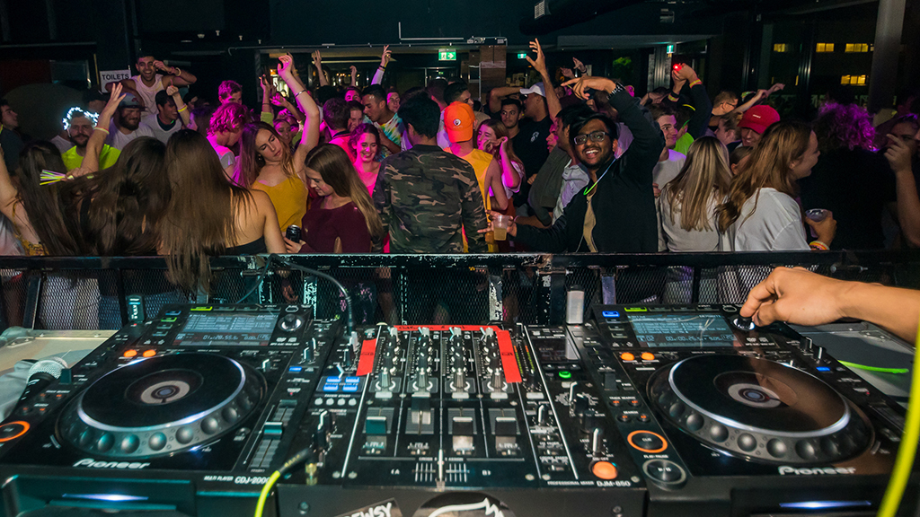 Photo taken from the stage of a DJ event showing DJ console and students dancing in the crowd.