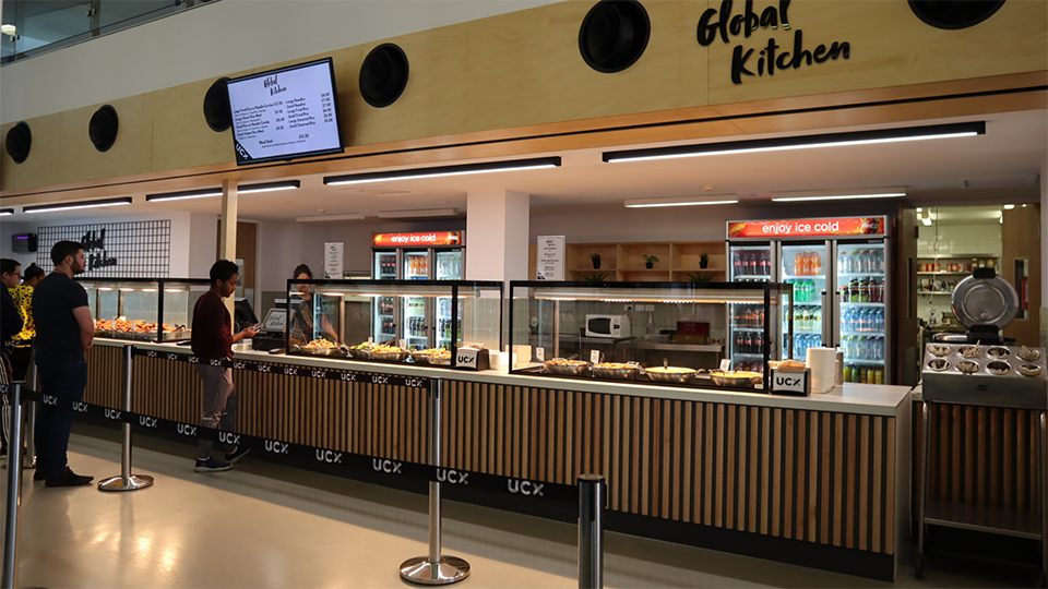 Global Kitchen shop front in the UC Refectory with UCX tensile barrier lines in front. Facade is wooden paneling, glass displays show the food options available and a TV screen mounted above the counter shows the menu. Two people are in the queue to order. 