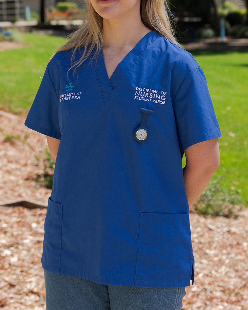Female wearing a dark blue University of Canberra branded scrub top. The top reads Discipline of Nursing Student Nurse. A medical fob watch is pinned to the scrub top underneath the logo on the left hand side. 