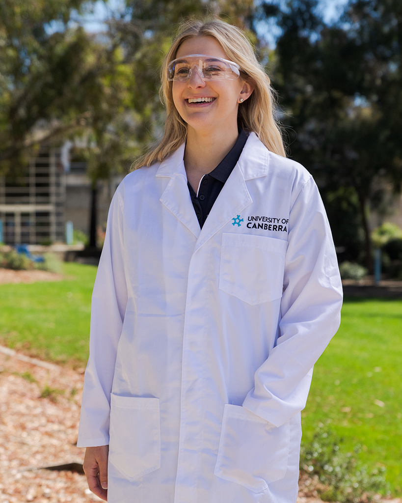 A young smiling female student wearing a white lab coat with a University of Canberra logo featuring over the left side chest pocket. She is also wearing clear safety glasses over her eyes