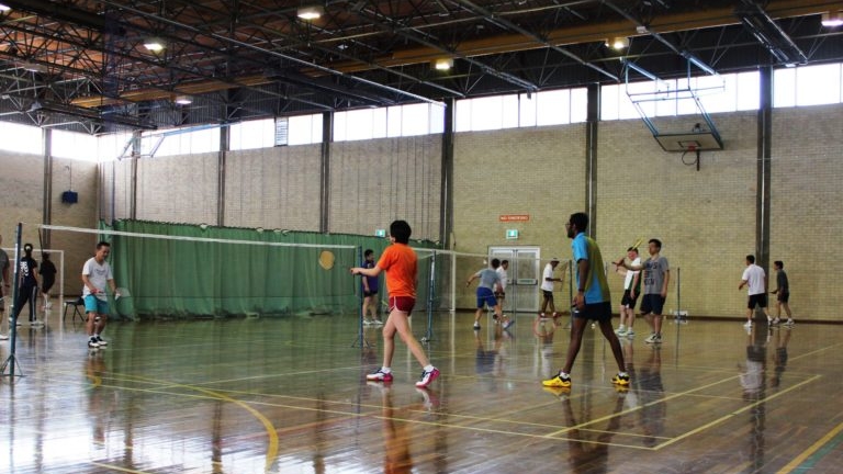 multiple people playing badminton on a polished timber floor with a badminton net