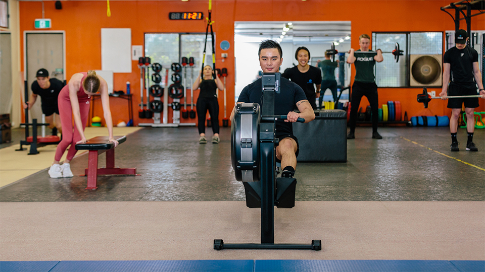 multiple people performing various gym exercises using a combination of body weight and free weights. In the foreground, a young man happily uses a row machine.