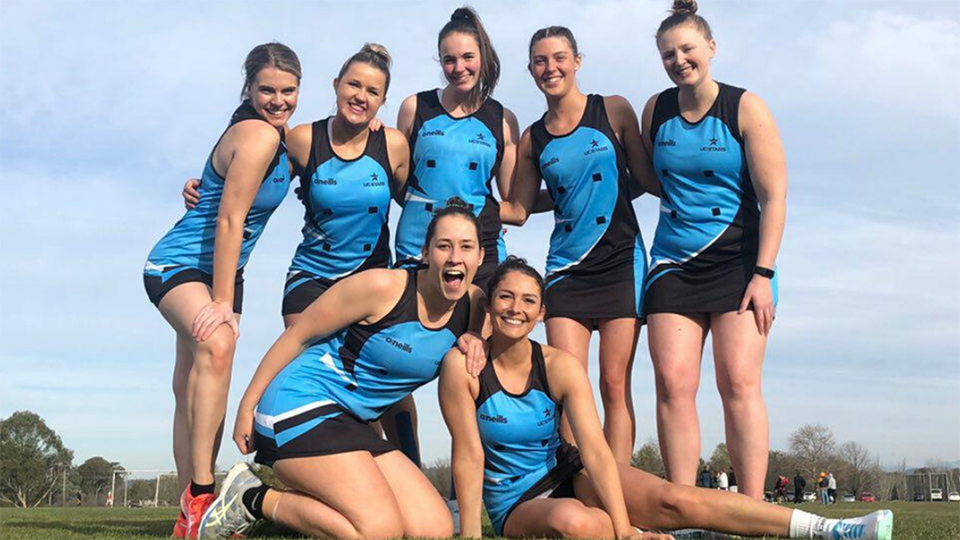 Group of seven female netball players posing for a team shot with 5 standing and 2 sitting on a green field. Players are wearing UC Stars blue uniforms and smiling.