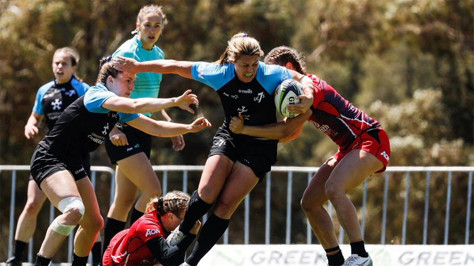 Determined female UC rugby 7's player in blue and black uniform running with ball, opposition player in red and black uniform trying to tackle UC player. Three other players and referee can be seen in background.