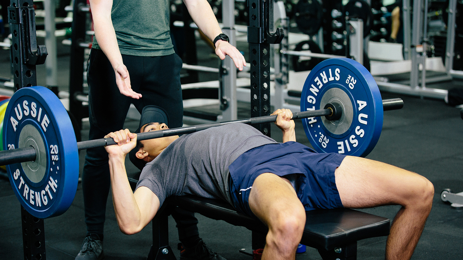 young man lying on a gym bench with a weighted bar across his chest. His elbows are bent as if to raise his arm and the bar. A man stands over him with hands outstretched ready to assist in raising the bar if required.